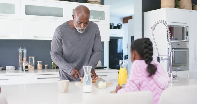 African American senior man at kitchen island slicing bread with knife, child watching making snack