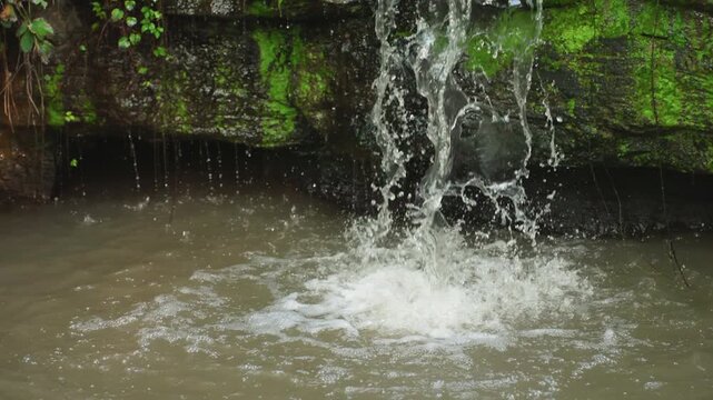 Closeup falling water into muddy pool, mosslined grotto with turbulent splash and rising sediment, wet stone ledge showing erosion, textured ripples and ambient damp atmosphere