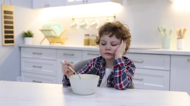 Unhappy little boy refusing to eat porridge at white marble table in kitchen