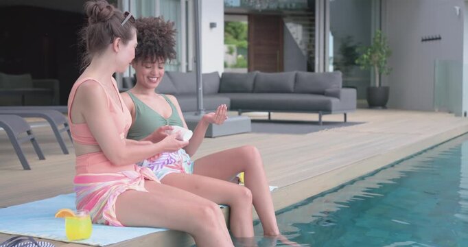 Diverse female friends relaxing on pool deck sharing sunscreen, protecting skin, applying lotion