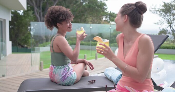 Diverse female friends toasting on pool deck in swimwear, clinking citrus cocktails, catching up