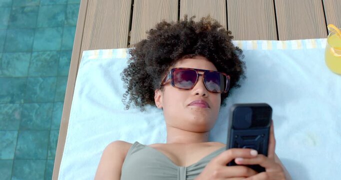 African American woman lying on towel on wooden pool deck, holding flip phone, checking messages