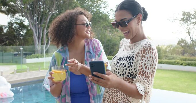 African American women standing by pool holding orange drinks while right woman sharing phone post