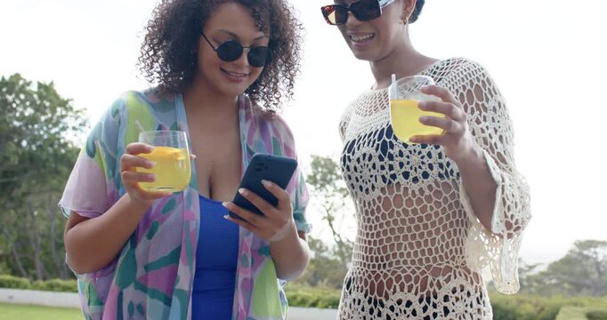 African American women at poolside holding drinks left scrolling smartphone right leaning laughing