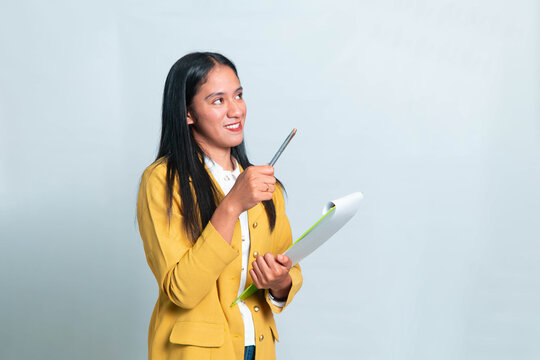 Smiling Indonesian woman wearing yellow blazer and white shirt, holding green clipboard and raising pen while looking up at copy space, isolated on white background.