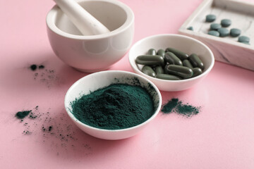 Bowls of spirulina capsules and powder with mortar on pink background