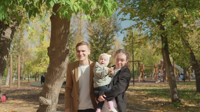 Family playing near big rope swing, parents hold and push baby while laughing, sunlit tree trunks and scattered leaves, warm autumn playground mood, protective gestures and joyful motion