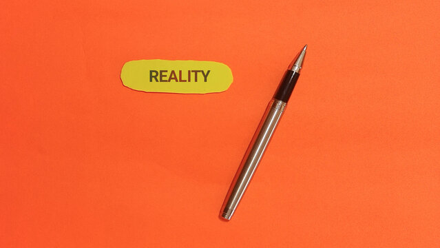 The word reality written on a yellow sticky note with a pen next to it against an orange background