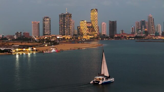 4K aerial view of a luxury yacht sailing near Colombo Port City beach with an illuminated modern skyline at dusk