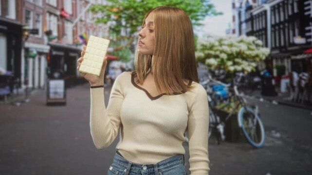Woman holding white chocolate bar with raised right hand on a busy street with bicycles and storefronts; indulgence craving.