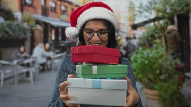 Woman wearing santa hat and glasses holding a stack of gift boxes on a busy city street; holiday cheer.