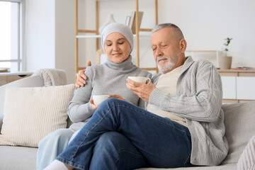 Mature woman after chemotherapy with her husband drinking tea on sofa at home