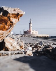 Fototapeta premium Iconic historic lighthouse standing on a harbor island with a vintage lilac facade and chocolate brown rocky foreground for travel tourism and souvenir posters