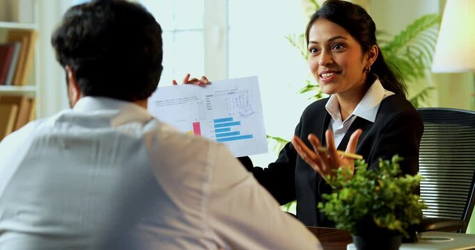 Indian young financial planner meeting young couple in modern office discussing on graph and reviewing growth reports across a table to ensure long-term wealth and investment success.