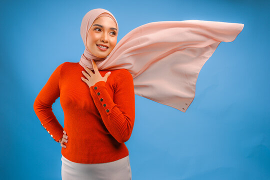 Elegant young Asian Muslim woman with hijab waving in the wind, modelling pose on blue background.