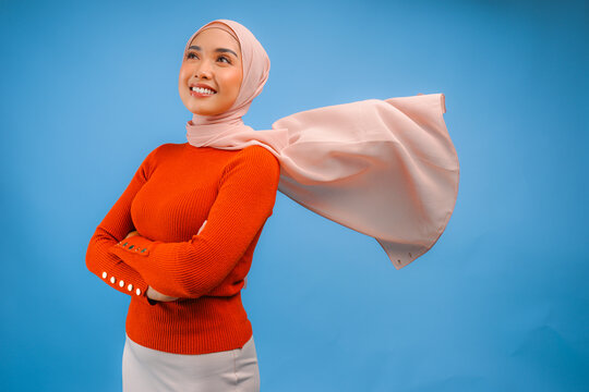 Elegant young Asian Muslim woman with hijab waving in the wind, modelling pose on blue background.