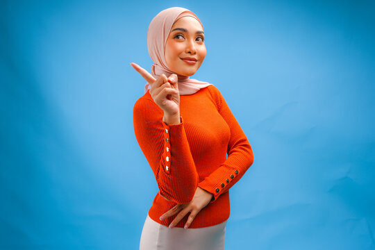 Happy young Asian Muslim woman wearing hijab pointing to the copy space behind her.
