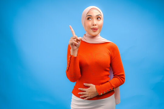 Hungry young Asian Muslim woman is holding her stomach while pointing to the copy space upwards.