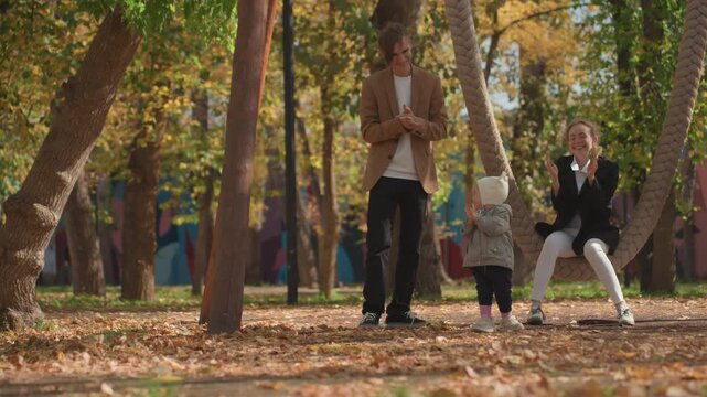 Toddler taking first steps with parents in sunlit autumn park, married couple support child near rope swing, leaves scattered underfoot, warm coats, gentle laughter and encouraging gestures capturing