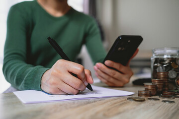 Fototapeta premium Woman writing budget notes while using smartphone with coins and savings jar on table. Representing personal finance, budgeting, saving money and financial planning.