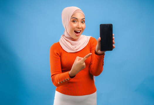 Pretty Asian Muslim woman in hijab smiling while showing the copy space on phone screen.