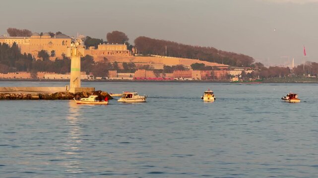 Scenic view of the Topkapi Palace and historic lighthouse on the Bosphorus coast at sunset. Small boats cruising the sea in Istanbul, Turkey. Iconic Ottoman landmark and maritime landscape.