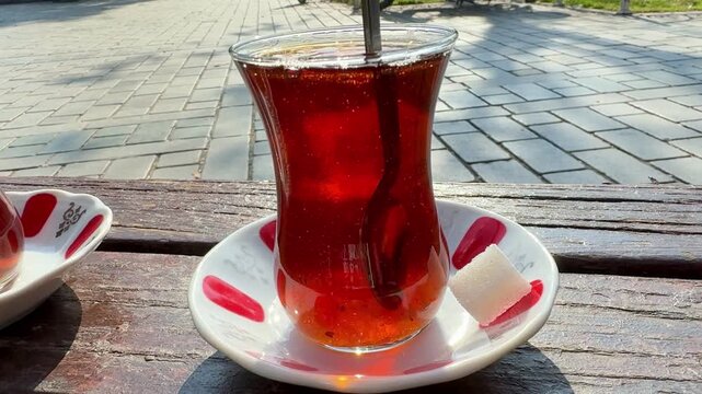 Traditional tulip-shaped glass of hot Turkish black tea on a red-patterned saucer. Authentic tea service in an outdoor Istanbul cafe with a sugar cube. Concept of hospitality and culture.