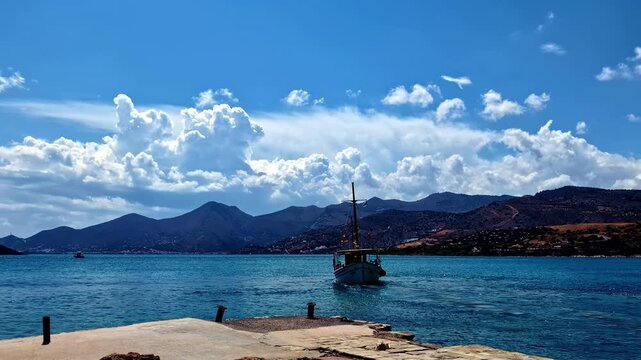 A small boat approaches the shore with mountains and Spinalonga island visible across the blue water, Crete, Greece