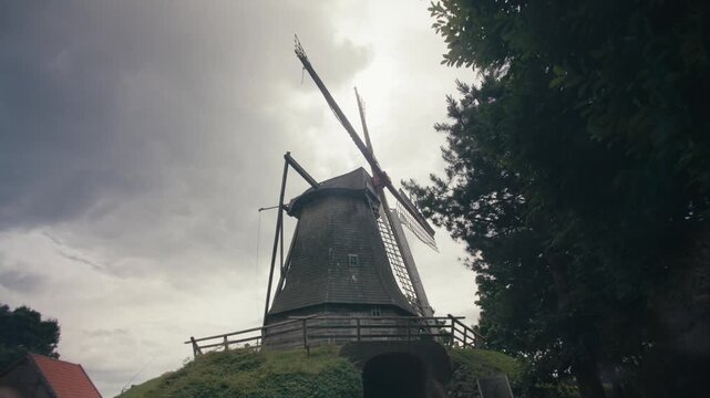 Low angle locked off shot of a historic wooden windmill on a hill against a dramatic cloudy sky. Dark moody atmosphere, surrounded by trees in northern Germany.