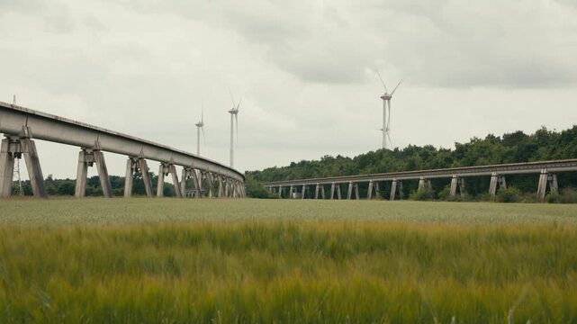 Locked off shot of the historic Transrapid maglev test track curving through a green wheat field near Lathen, Germany, with wind turbines in the background.