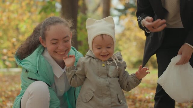 White parents helping toddler take first steps among golden autumn leaves, father supporting, mother encouraging with kisses and smiles, seeds and pigeons nearby, joyful family milestone