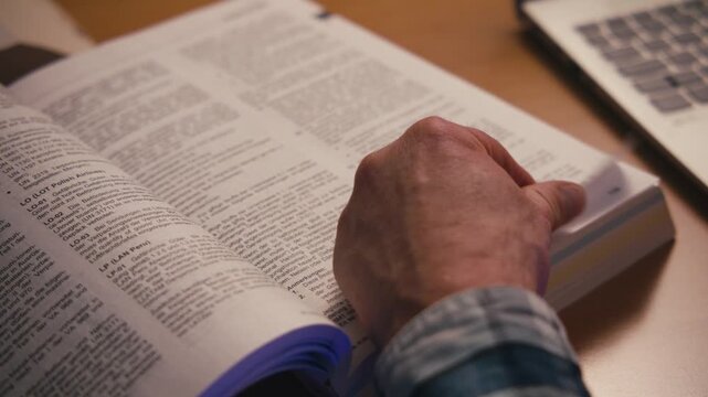 Close-up handheld shot of a man's hand turning pages of a large German reference book at a desk. Regulations text visible, calculator in background.