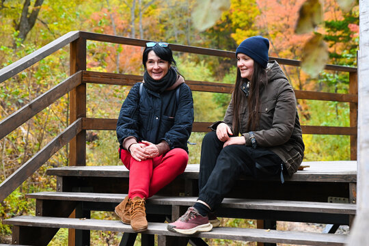 Two Russian women travelers friends laughing and talking while sitting on wooden stairs in autumn forest Russia one 50 years old and one 35 years old
