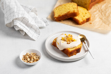 Orange Zest Cream-Topped Cake Slice with Nuts. A golden sponge cake slice topped with a smooth white frosting, garnished with bright orange zest and chopped nuts. Plated on a white round dish with a g