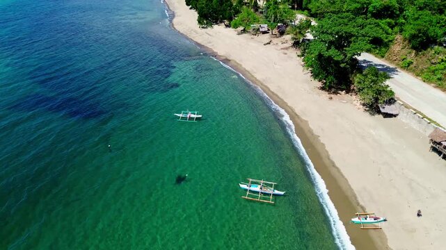 Aeral drone dolly up fly over of a serene tropical beach with outrigger boats and lush greenery along the shore adjacent to a coastal road with a person walking toward the sea in Basay, Philippines