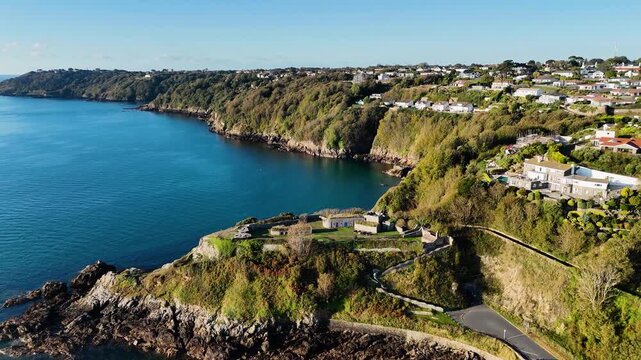 Flight over Clarence Battery Guernsey towards Fort George with beautiful wooded cliffs and calm deep blue sea on sunny day with cloudless sky