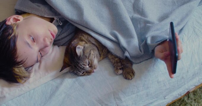 A teenage girl uses her smartphone while lying in bed, her cat resting peacefully next to her, both enjoying a quiet and relaxed moment together. 60 fps 4K footage.