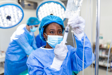 Focused black female anesthesiologist adjusting IV drip with syringe in operating room. Healthcare and medicine concept. Medical professional in mask and scrubs preparing anesthesia for surgery.