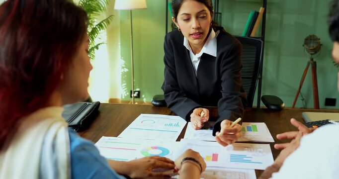 Indian young financial planner meeting young couple in modern office discussing on graph and reviewing multiple reports spread over the table to plan their future investment strategy.