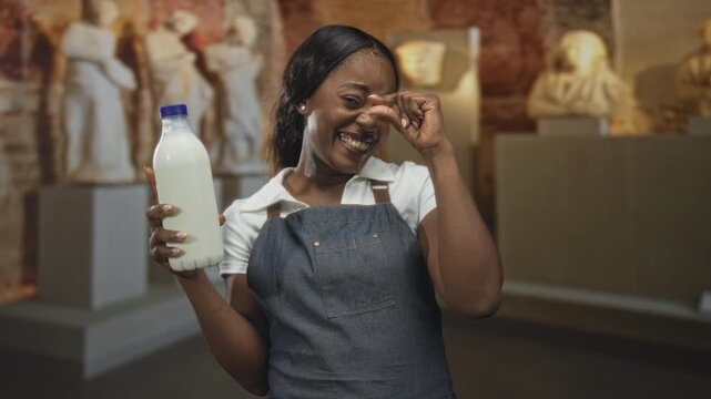 Woman in apron holds a milk bottle in one hand and pinches her nose between thumb and finger while smiling amid statue pedestals in a building; amused curiosity.