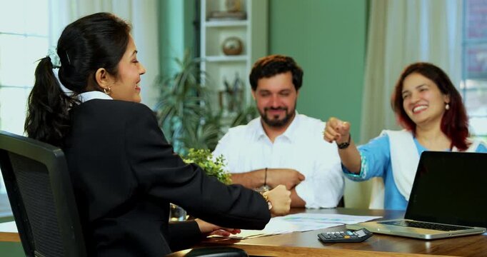 Indian professional executive meeting young couple in modern office shaking hands and showing thumbs up to camera while sitting across a table to confirm a successful financial agreement.