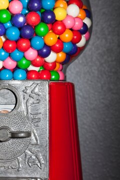 Tightly framed image of colorful gumballs in a red vintage gumball machine with coin slot, set against a textured gray wall.