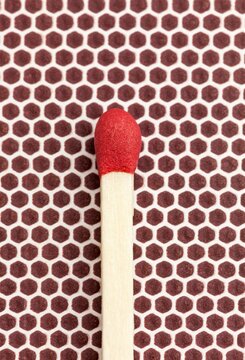 Macro photo of a single red-tipped wooden matchstick resting on the brown textured flint striking surface of a matchbox.