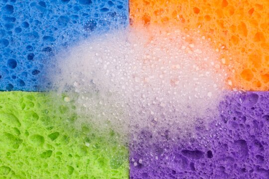 Four brightly colored household sponges arranged in a grid formation with a mound of soap bubbles in the center. Vivid colors - blue, orange, green, and purple