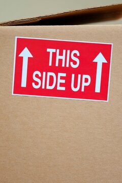 Close-up of a red "This Side Up" sticker with white arrows on a cardboard shipping box, handling fragile packages during transit and delivery.