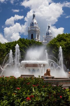 Scenic view of the Fountain of the Lions with the twin towers of the historic Ponce Cathedral rising above lush greenery and flowering shrubs under a partly cloudy sky in Ponce, Puerto Rico.