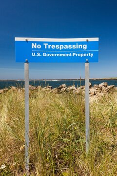 Blue "No Trespassing, U.S. Government Property" sign posted in grassy dunes near the shoreline in Winchester Bay
