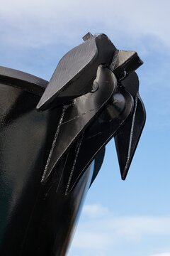 Close-up of a large black anchor mounted on the bow of a ship, the sharp lines and glossy finish of the anchor stand out against a bright blue sky.