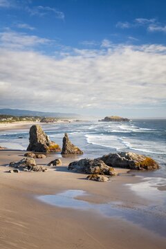 Dramatic sea stacks rise from the sandy shoreline of Bandon Beach, Oregon, with waves rolling in under a mix of blue skies and clouds on a bright coastal day.