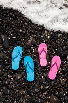 Two pairs of wet flip flops, one blue and one pink, rest on a rocky shoreline in Florence, Oregon as ocean foam approaches.
